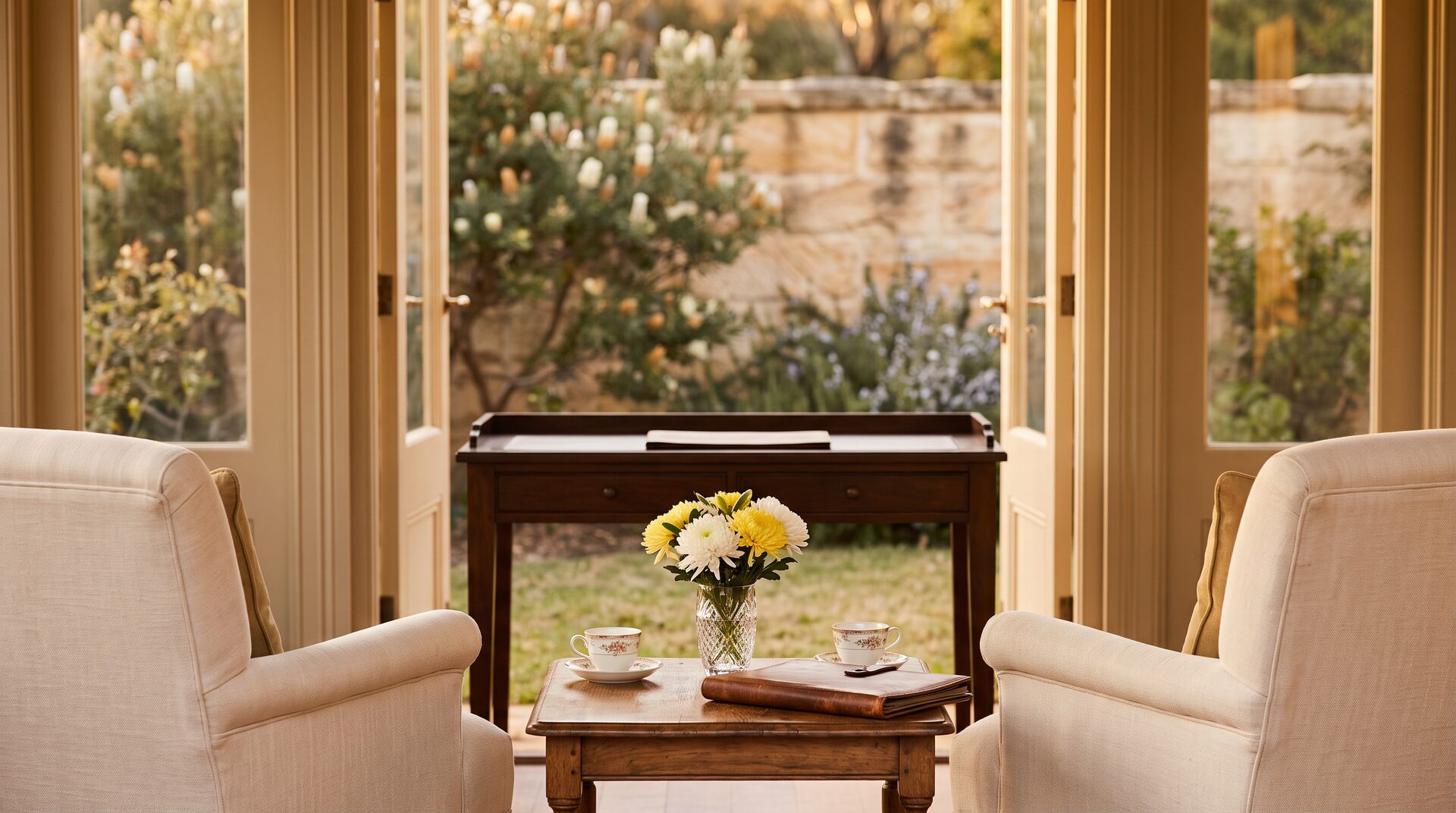A private Australian study with two cream armchairs, tea cups, chrysanthemums, a leather portfolio and open French doors to a sandstone-walled garden.